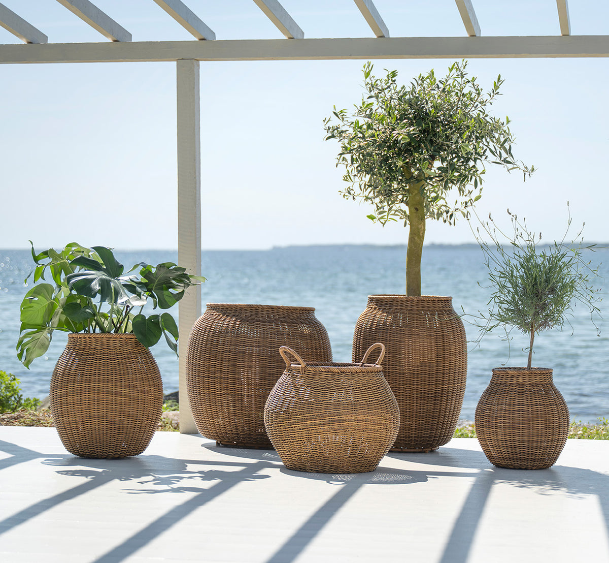 This is a lifestyle image of Alma Basket in chestnut core weave ArtFibre Aluminium, outdoors on a sun-lit patio with a white wall and tile flooring.