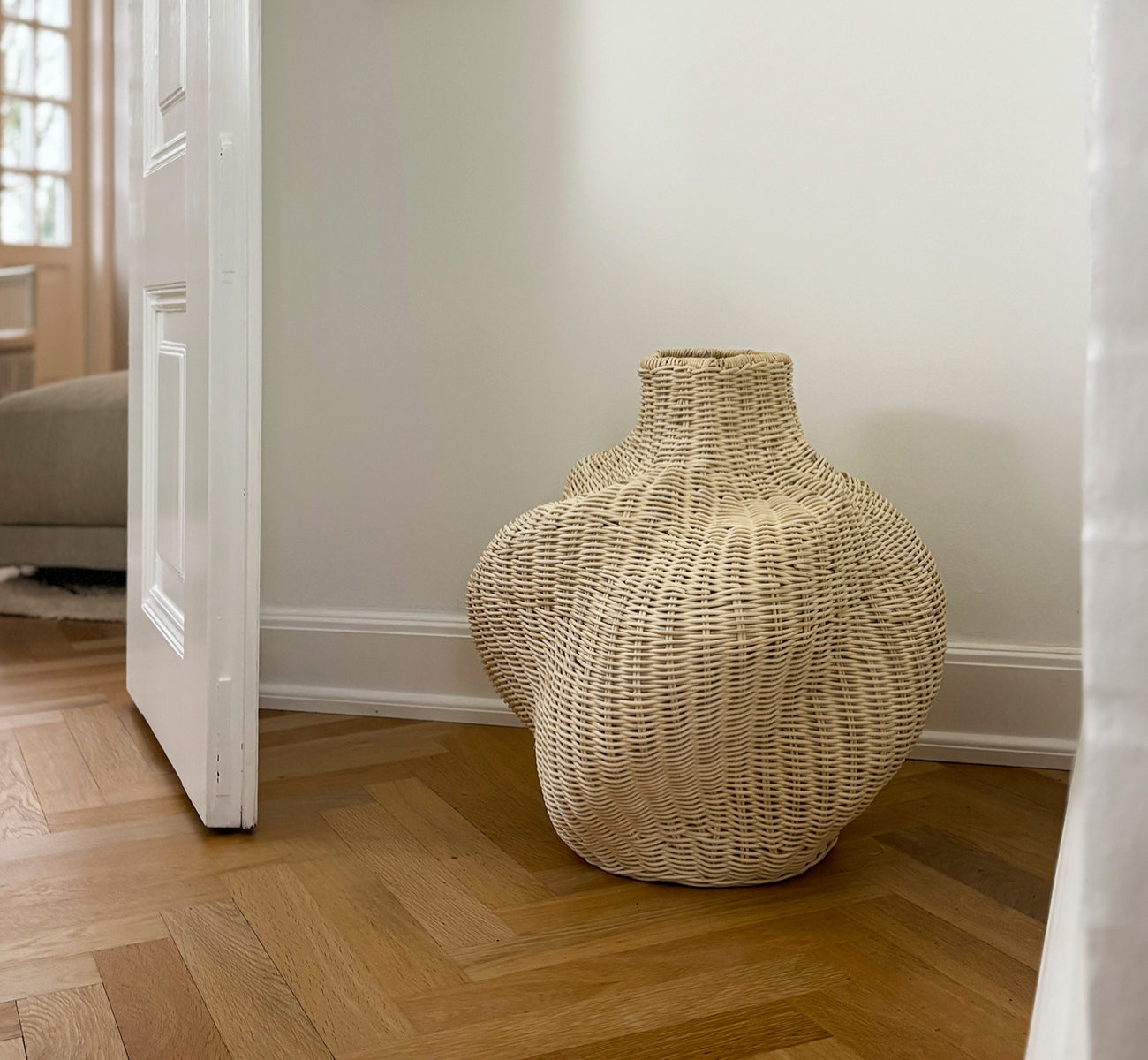 This is a lifestyle image of Solaris Basket Ø40 cm in natural core weave iron frame. Styled with dried flowers by a white door on parquet flooring.