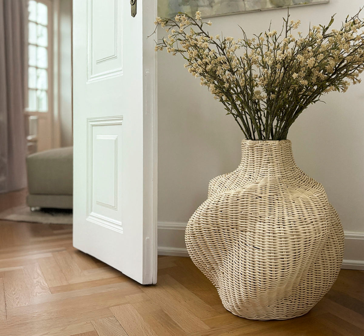 This is a lifestyle image of Solaris Basket Ø46 cm in natural core weave iron frame. Styled with dried flowers beside a white door on a parquet floor.