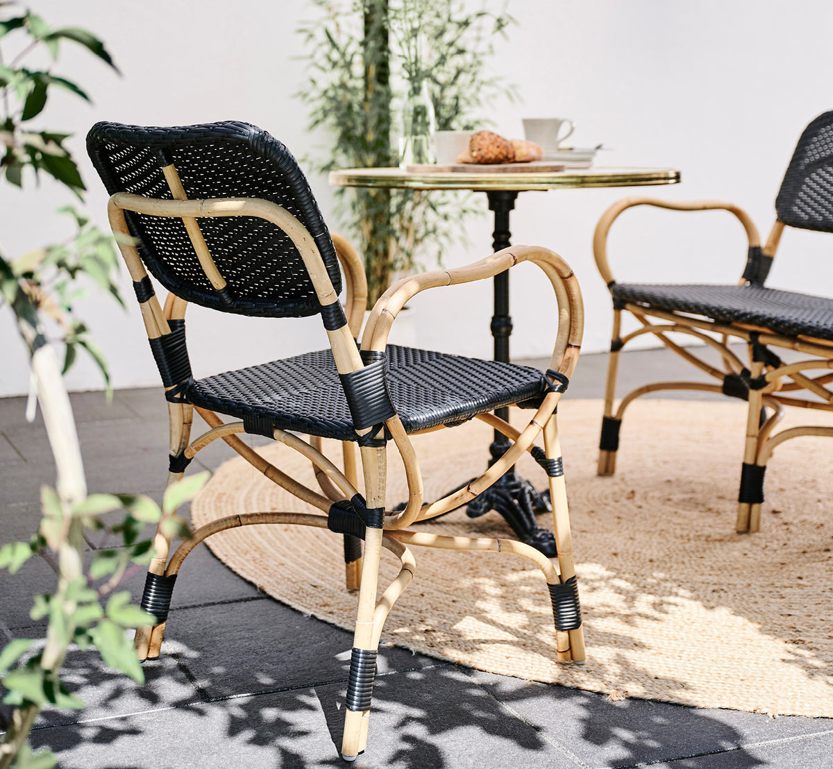 This is a lifestyle image of Bistro Dining Chair in skin on Rattan frame, in an outdoor café setting with a bistro table on dark tile flooring.