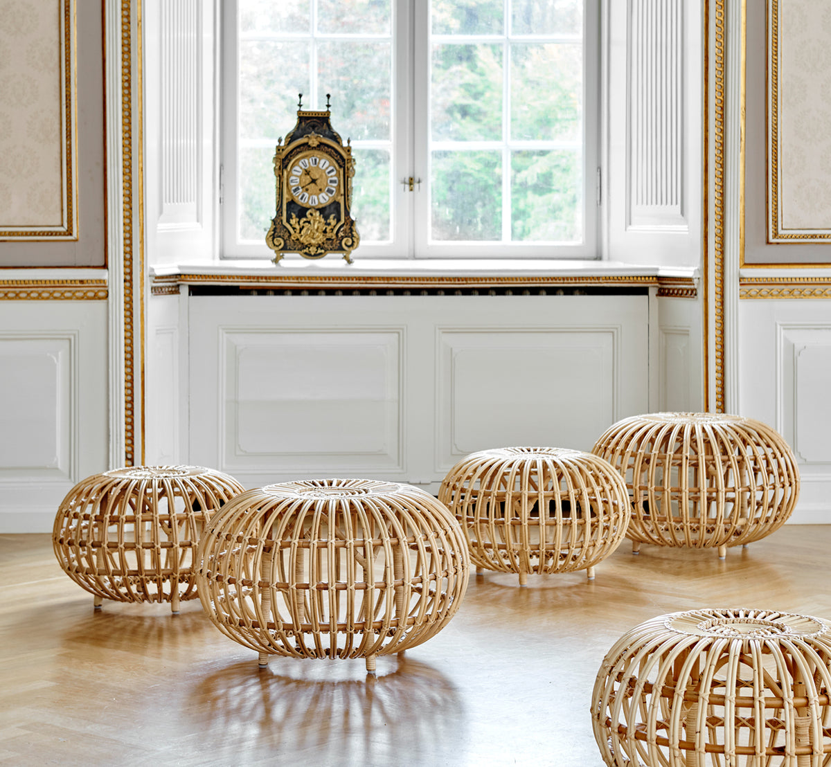 This is a lifestyle image of Ottoman Small in natural skin on rattan. Clustered in a classical panelled interior with an antique clock.