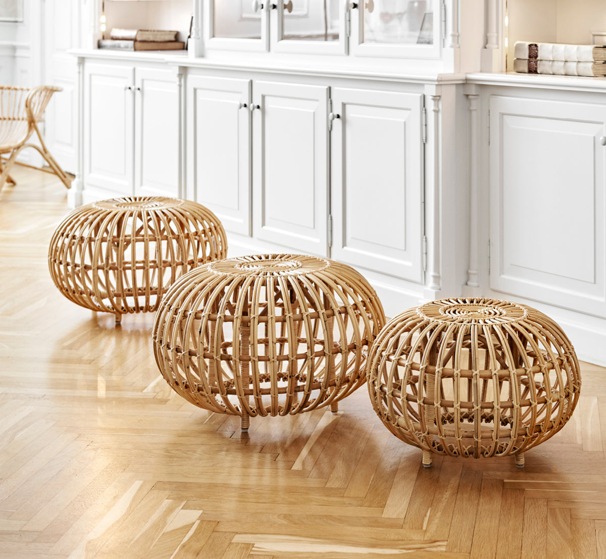 This is a lifestyle image of Ottoman Small in natural skin on rattan. Clustered in a classical panelled interior with an antique clock.