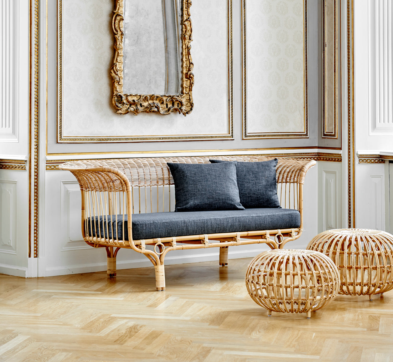 This is a lifestyle image of Ottoman Small in natural skin on rattan. Clustered in a classical panelled interior with an antique clock.