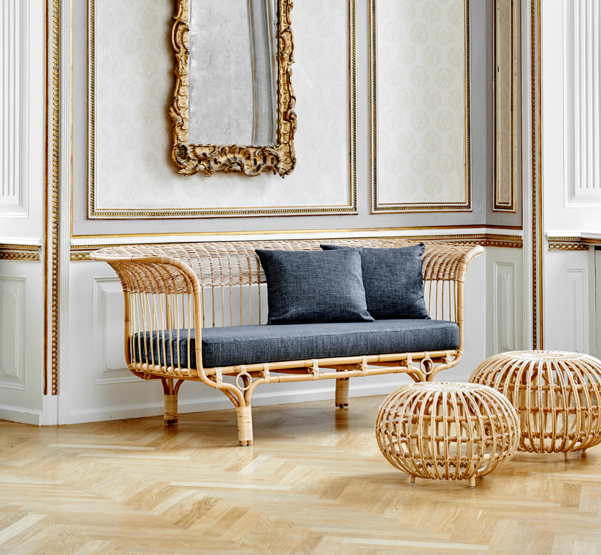 This is a lifestyle image of Ottoman Small in natural skin on rattan. Clustered in a classical panelled interior with an antique clock.