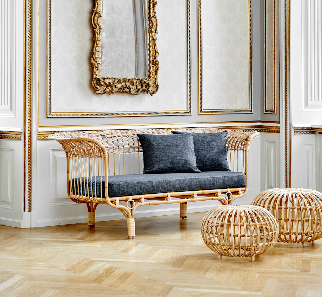 This is a lifestyle image of Ottoman Large in natural skin on rattan. Clustered in a classical panelled interior with a gilded clock.