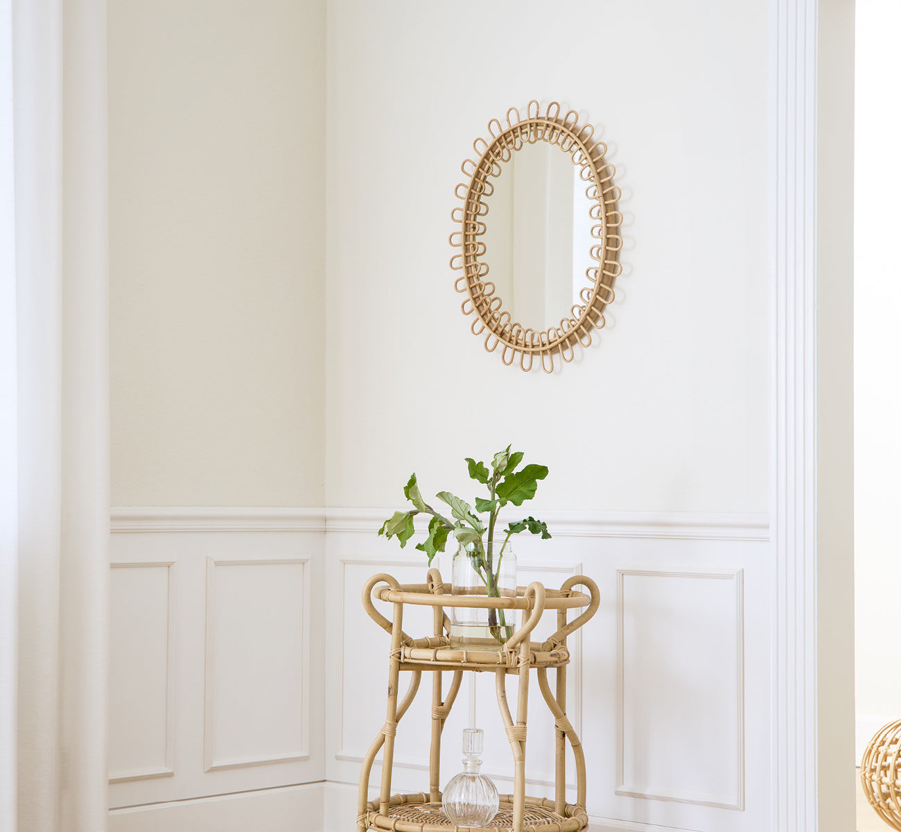 This is a lifestyle image of Luella Mirror in polished natural rattan. Mounted on a white paneled wall above a rattan bar cart with greenery.