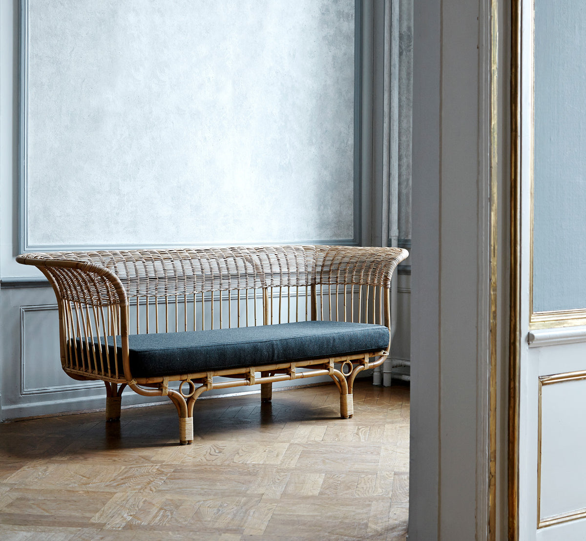 This is a lifestyle image of Belladonna Sofa in polished natural rattan, in a bright Scandinavian interior seen through a white doorway.