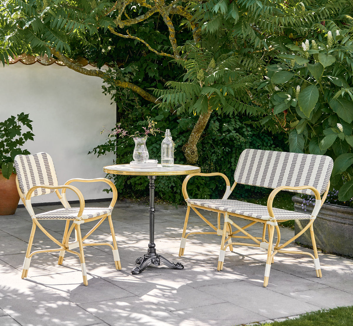 This is a lifestyle image of Bistro Exterior Chair in natural Aluminium ArtFibre, in an outdoor garden café with a bistro table and surrounding trees.