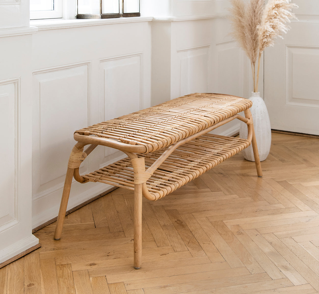This is a lifestyle image of Machiya Bench in natural skin on rattan. White paneled hallway with herringbone floor and pampas grass vase.