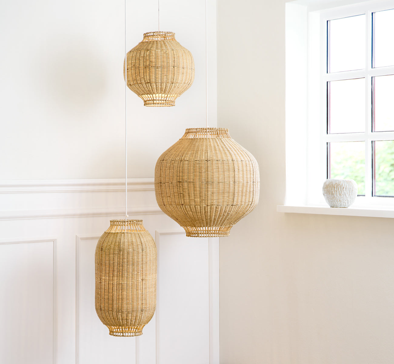 This is a lifestyle image of Kou Lampshade in natural core weave rattan frame. Three sizes hang together in a bright white paneled room.