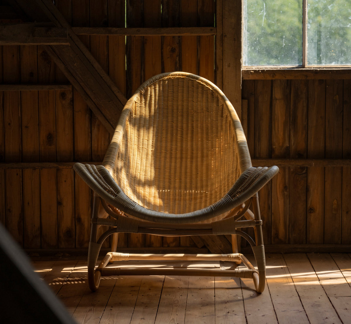 This is a lifestyle image of Pacifique Lounge Chair in natural core weave rattan frame. In a wooden barn with warm window light.