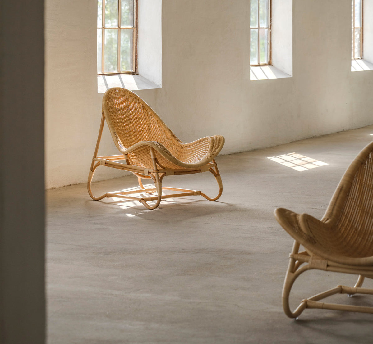 This is a lifestyle image of Pacifique Lounge Chair in natural core weave rattan frame. Pair in a sunlit room with concrete floor.