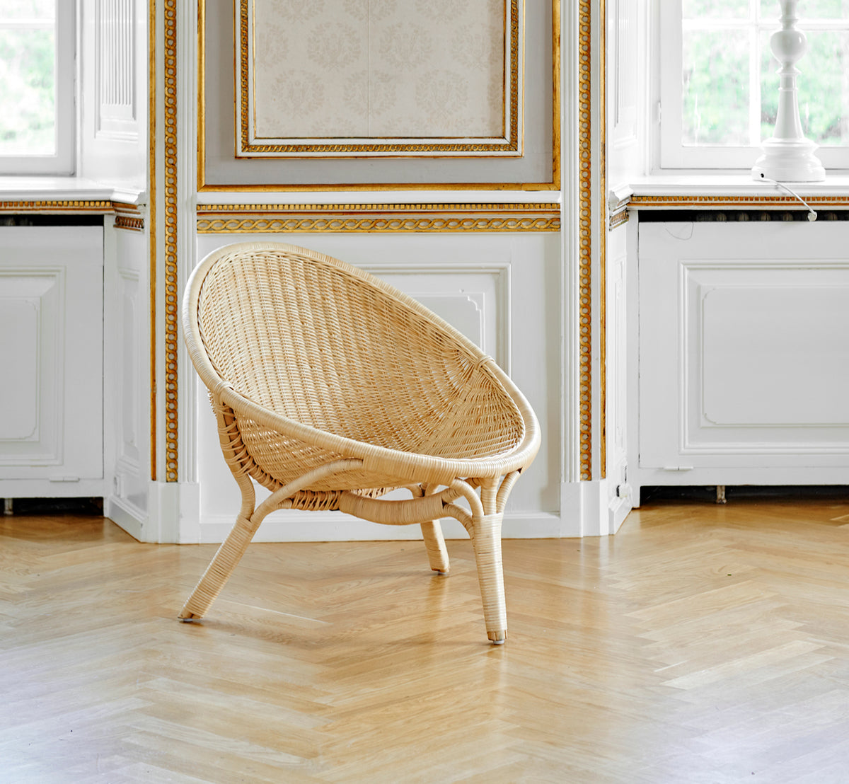 This is a lifestyle image of Rana Lounge Chair in natural core weave rattan frame. Standing alone in a room with gilt panelling and parquet flooring.