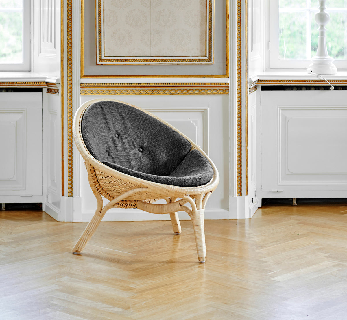 This is a lifestyle image of Rana Lounge Chair in natural core weave rattan frame. Standing alone in a room with gilt panelling and parquet flooring.