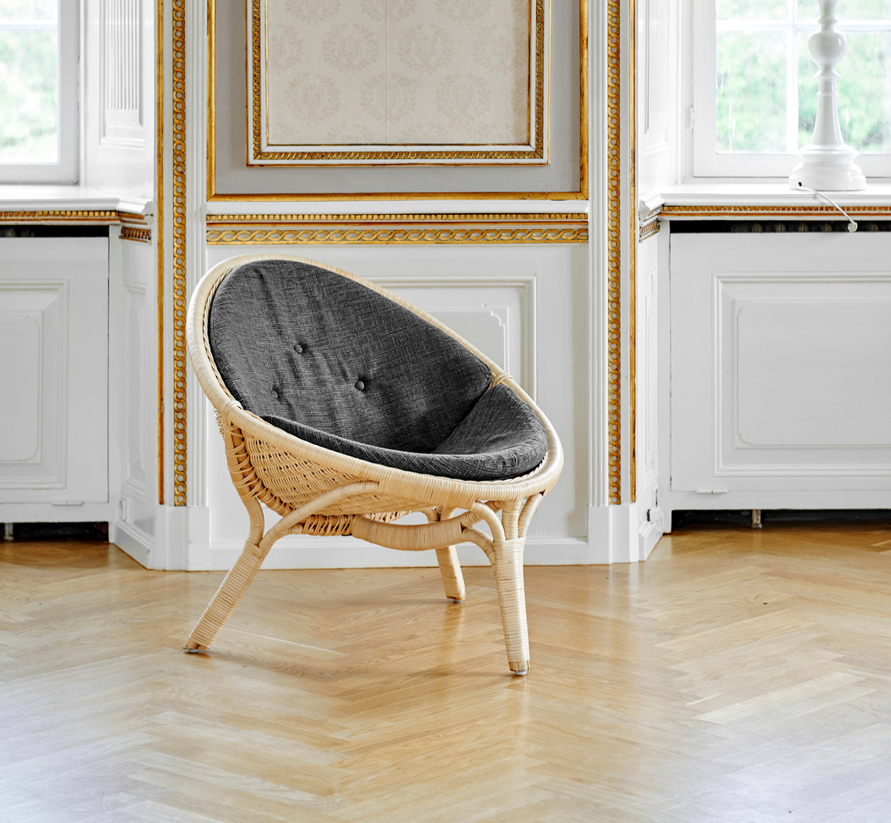 This is a lifestyle image of Rana Lounge Chair in natural core weave rattan frame. Standing alone in a room with gilt panelling and parquet flooring.