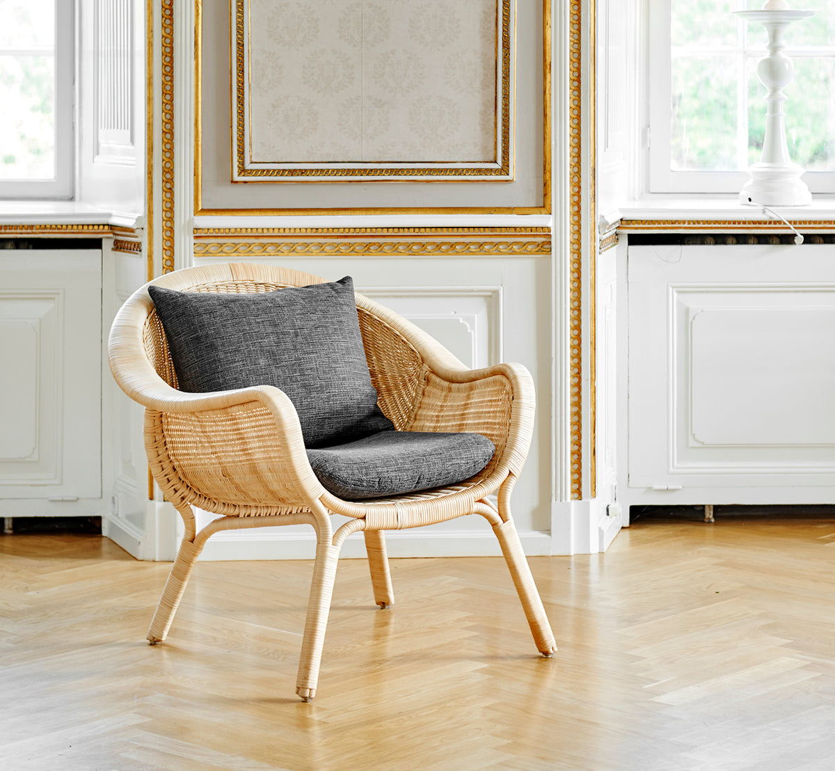 This is a lifestyle image of Madame Lounge Chair in natural core weave rattan frame. Elegant gilded room with oak floor and grey cushion.
