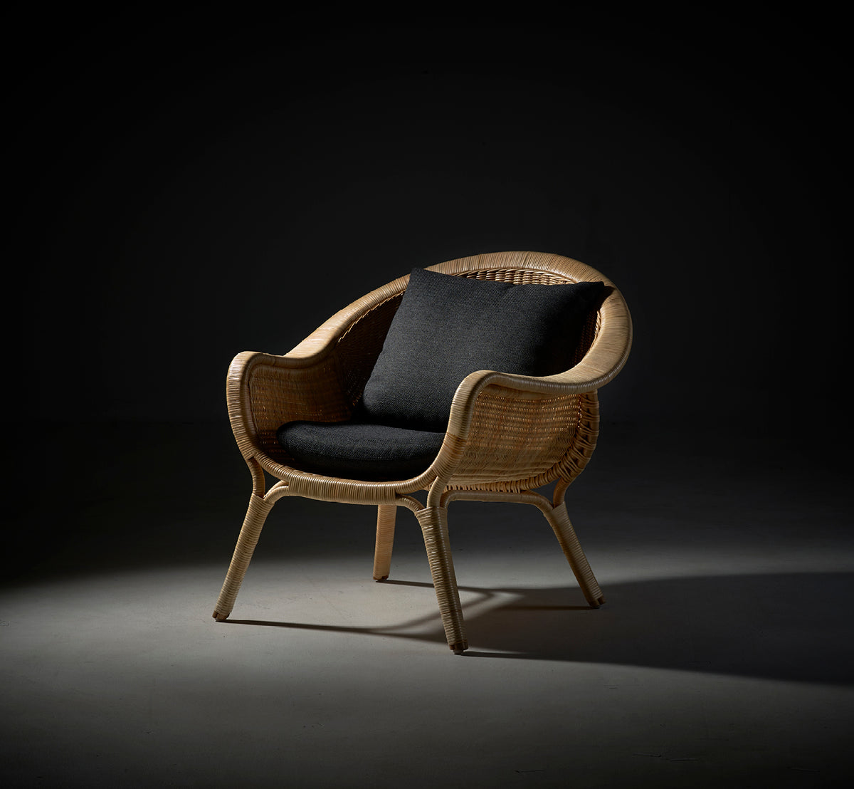 This is a lifestyle image of Madame Lounge Chair in natural core weave rattan frame. Dramatic studio lighting with dark cushion on grey backdrop.