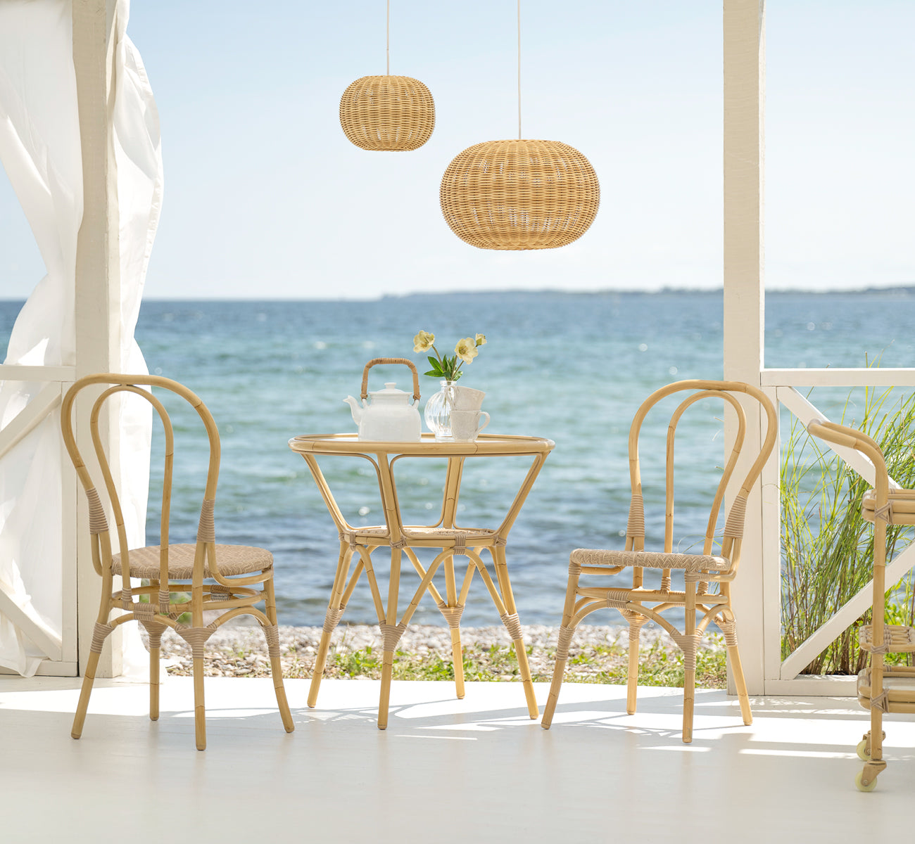 This is a lifestyle image of Lulu Exterior Dining Chair in natural aluminium artfibre. At a seaside terrace with a round table and ocean view.