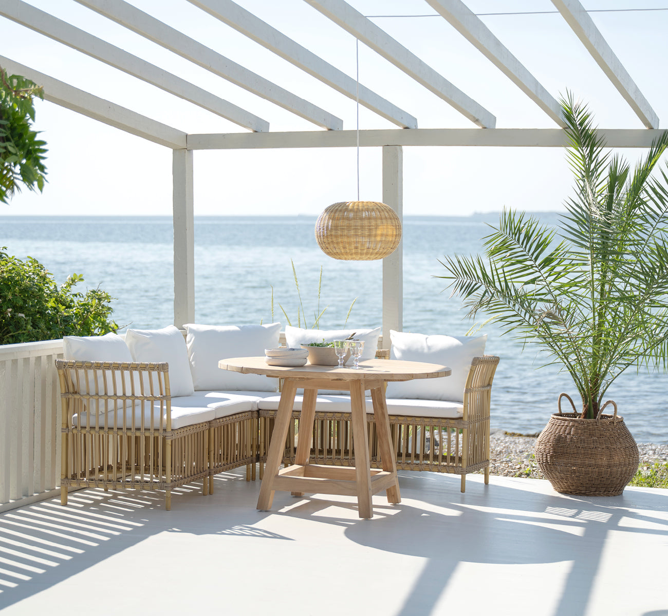 This is a lifestyle image of Meghan Exterior Dining Bench Corner Module in natural aluminium artfibre. With round table by the sea.