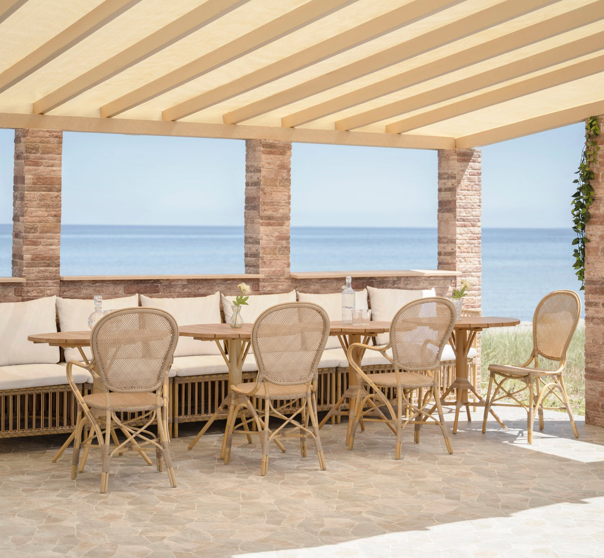 This is a lifestyle image of Meghan Exterior Dining Bench-Center Module in natural aluminium artfibre. Under a pergola by the sea.