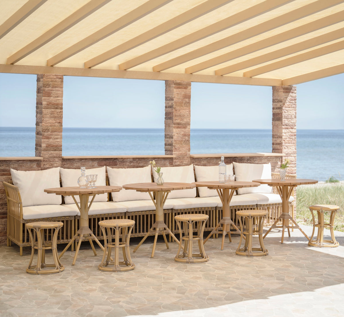 This is a lifestyle image of Meghan Exterior Dining Bench-Center Module in natural aluminium artfibre. Under a pergola by the sea.