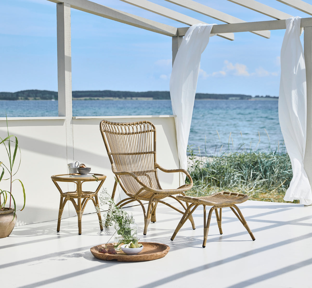 This is a lifestyle image of Monet Exterior Footstool in antique aluminium artfibre. On a beach deck with lounge chair and sea view.