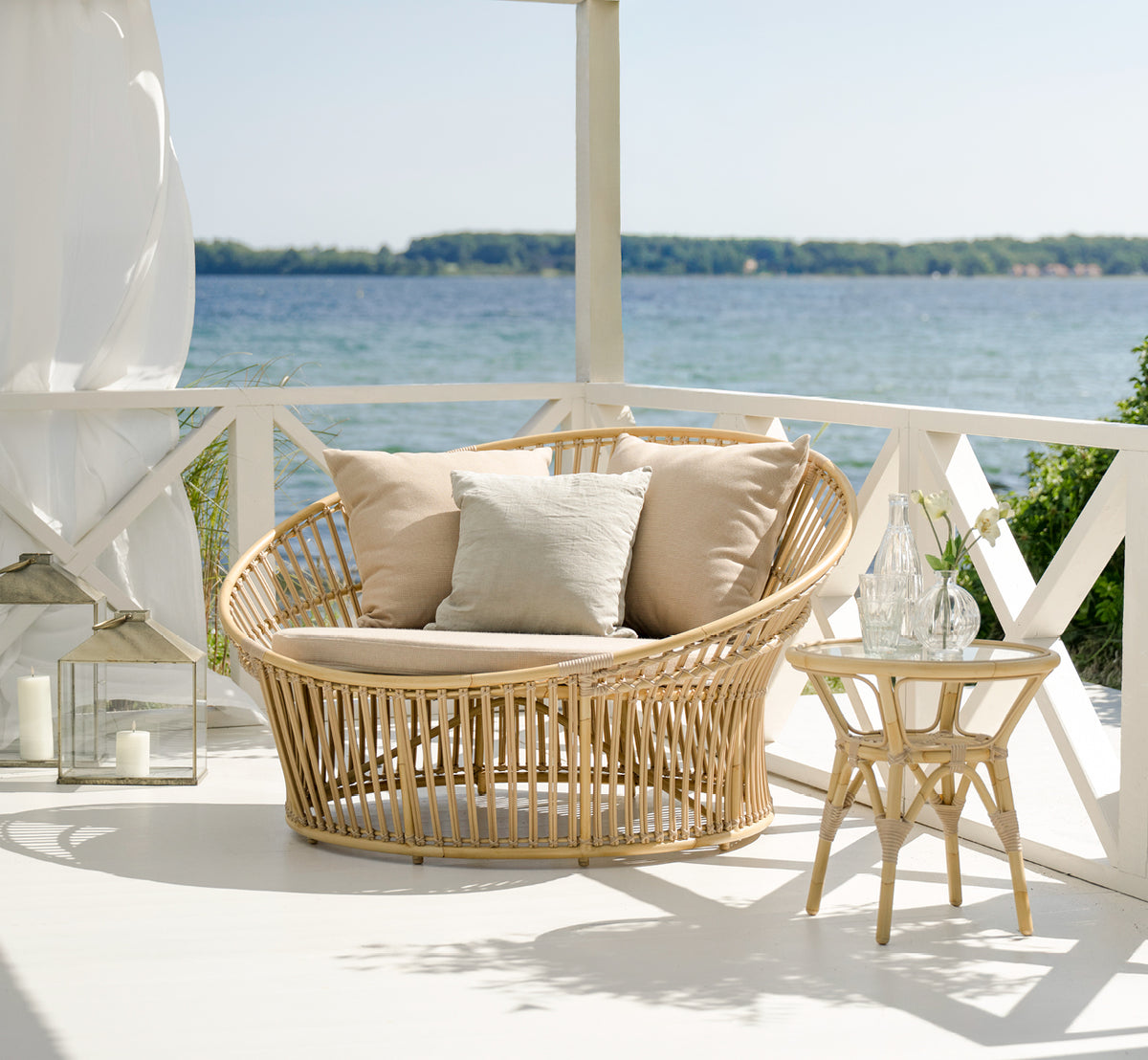 This is a lifestyle image of Tony Exterior Side Table in natural aluminium artfibre. On a wooden deck with jute rug by the sea.