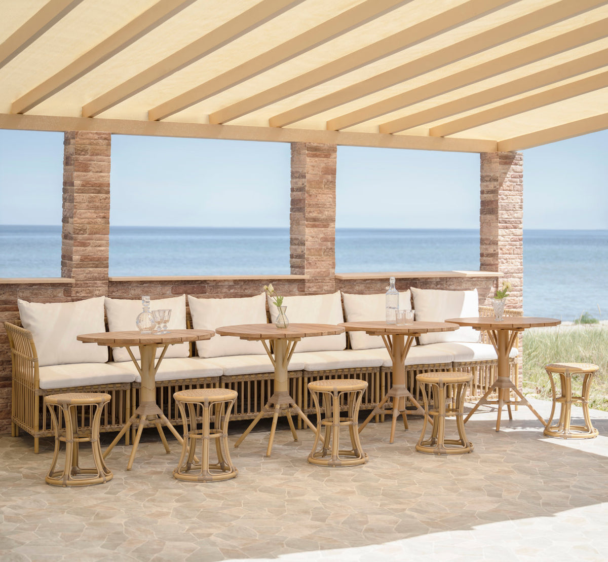 This is a lifestyle image of Nicole Cáfe Table-Teak Ø80 cm in natural aluminium artfibre. Bench with stools and café tables under a pergola.