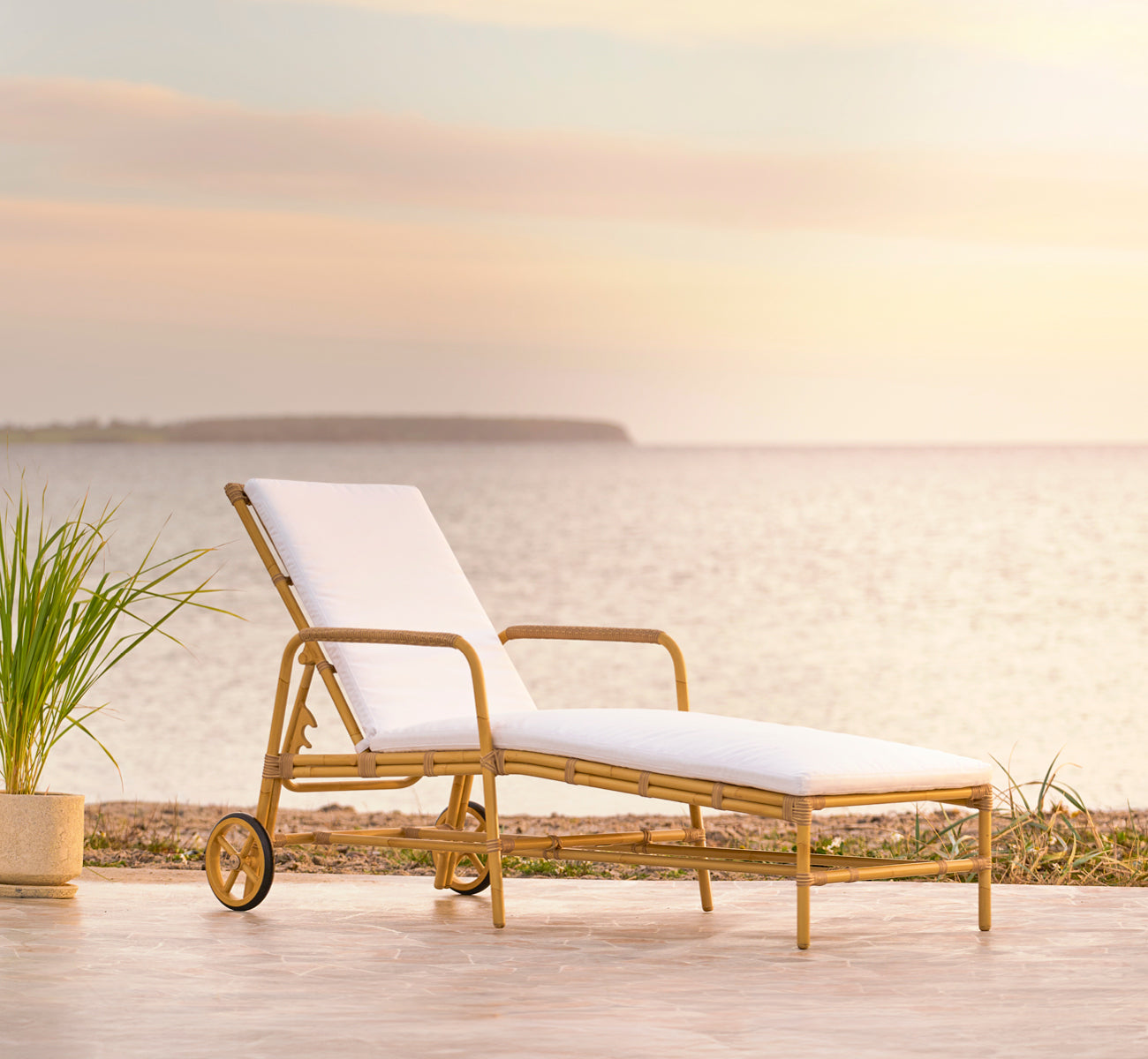 This is a lifestyle image of Josephine sunbed in natural aluminium artfibre, on a coastal stone terrace at sunset with sea views and beach grasses.