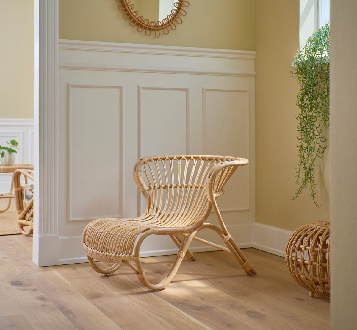 This is a lifestyle image of Fox Lounge Chair in polished natural rattan. By white panels with a rattan mirror.