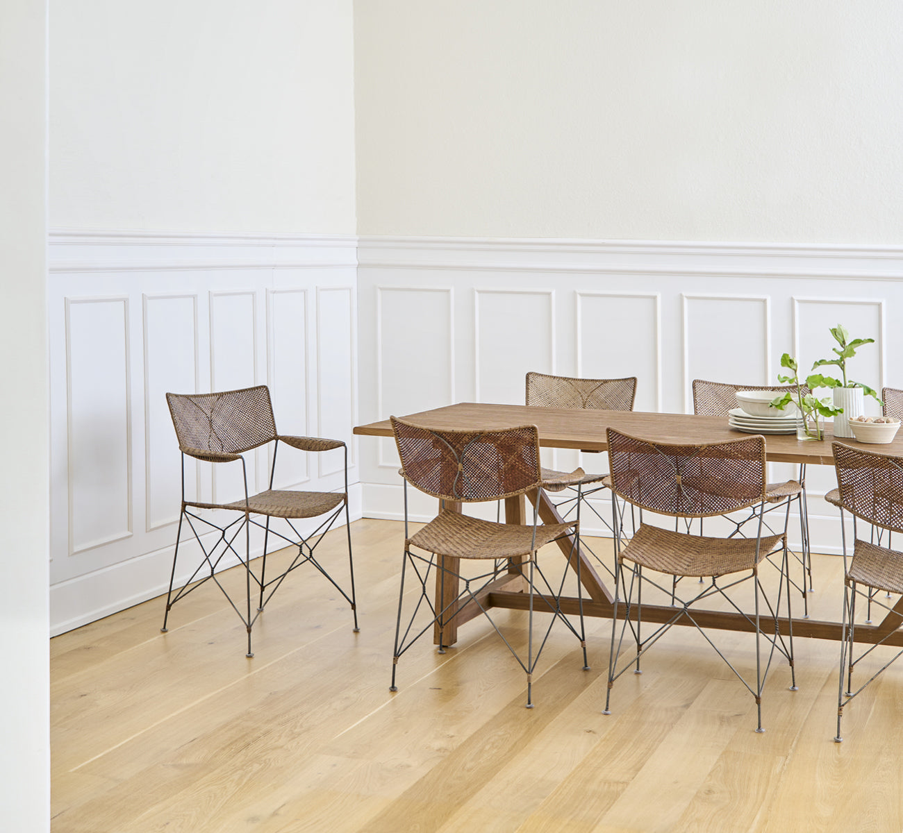 This is a lifestyle image of Rap Dining Chair in hazelnut rattan iron frame, in a bright interior with white panelled walls and herringbone parquet.