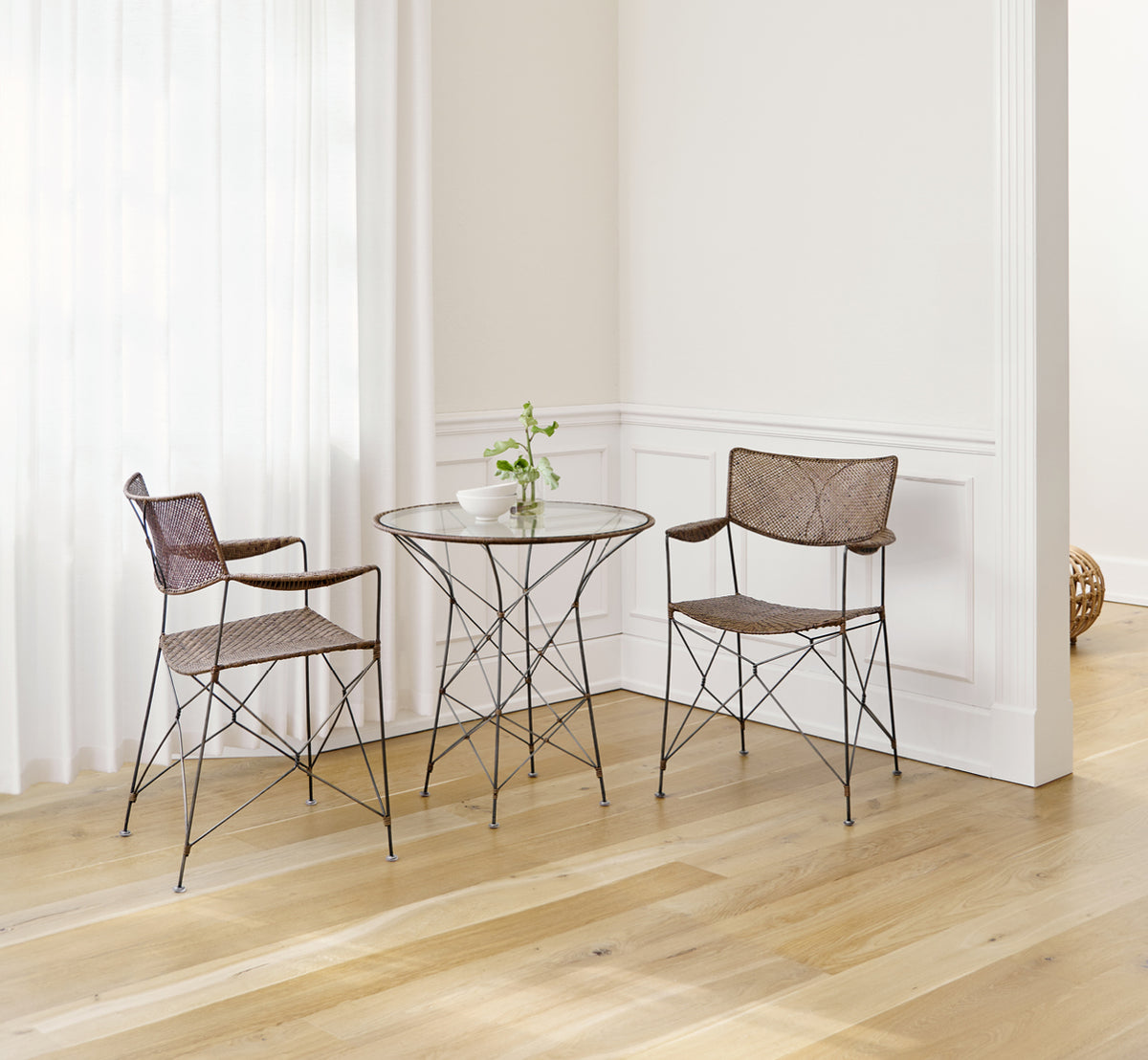 This is a lifestyle image of Rap Dining Chair in hazelnut rattan iron frame, in a bright interior with white panelled walls and herringbone parquet.