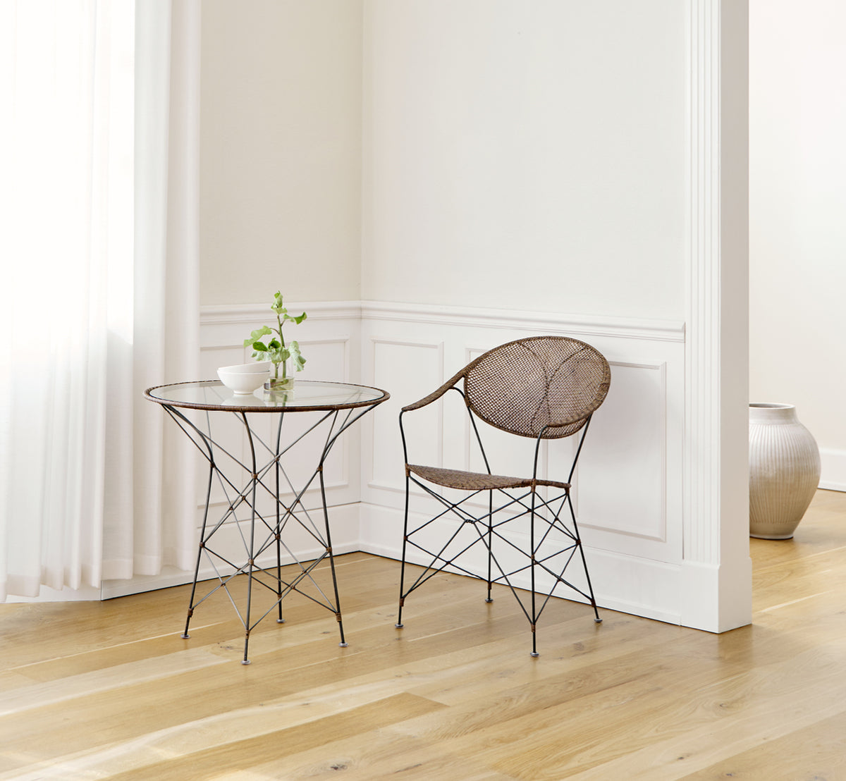 This is a lifestyle image of Funky Dining Chair in hazelnut rattan iron frame, in a bright interior with white panelled walls and herringbone parquet.